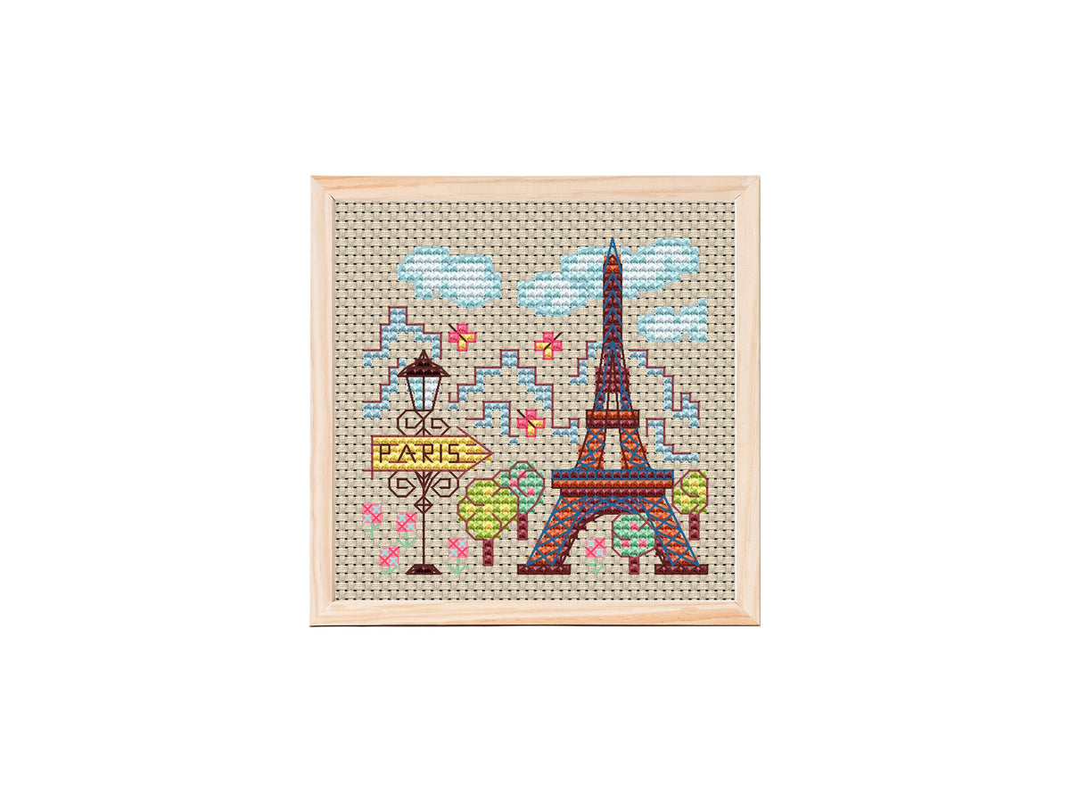 Cross stitch design of a small Paris cityscape showing a colorful Eiffel Tower, streetlamp with Paris sign, trees, clouds, and flowers on beige fabric.