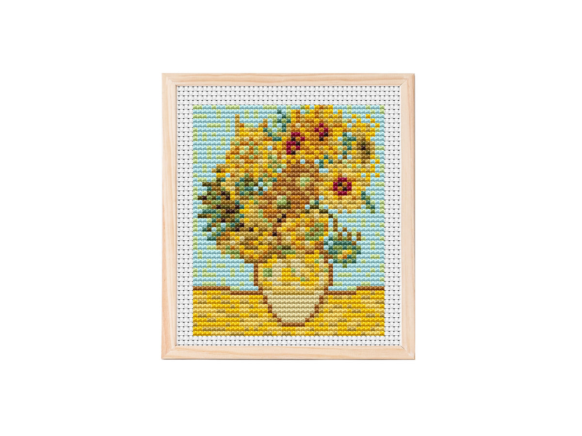 Cross stitch pattern of a vase filled with golden sunflowers on a yellow tabletop against a light blue background, stitched on white aida fabric.