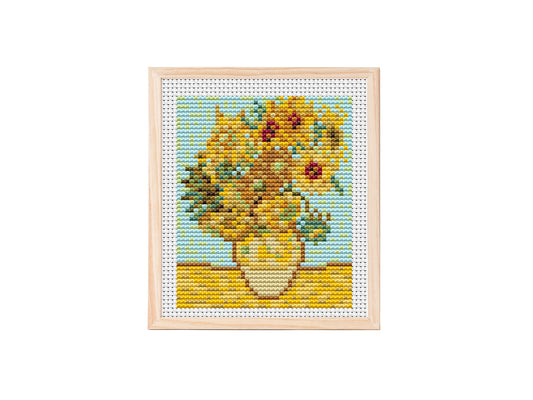 Cross stitch pattern of a vase filled with golden sunflowers on a yellow tabletop against a light blue background, stitched on white aida fabric.