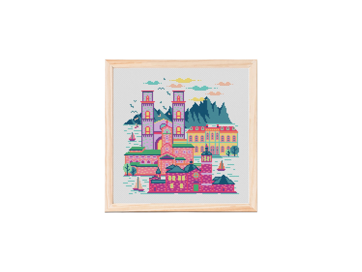 Colorful Montenegro-inspired cross stitch pattern showing a coastal cityscape with tall towers, domed buildings, sailboats on the water and dark mountains in the background on white fabric.